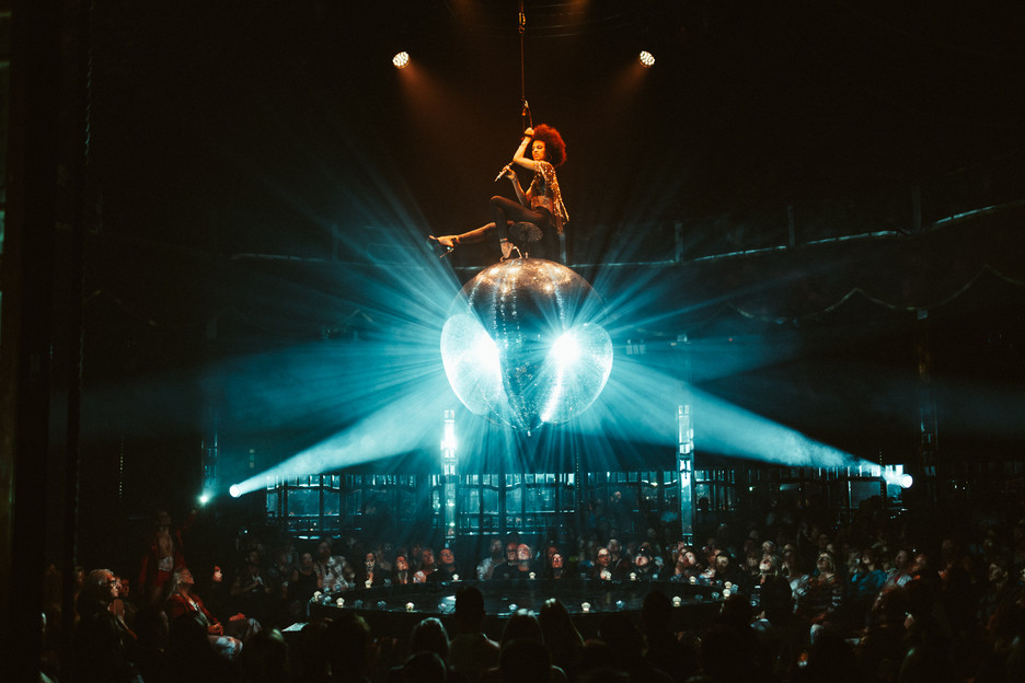 An aerialist performing above a glowing mirror ball over a darkened theatre audience
