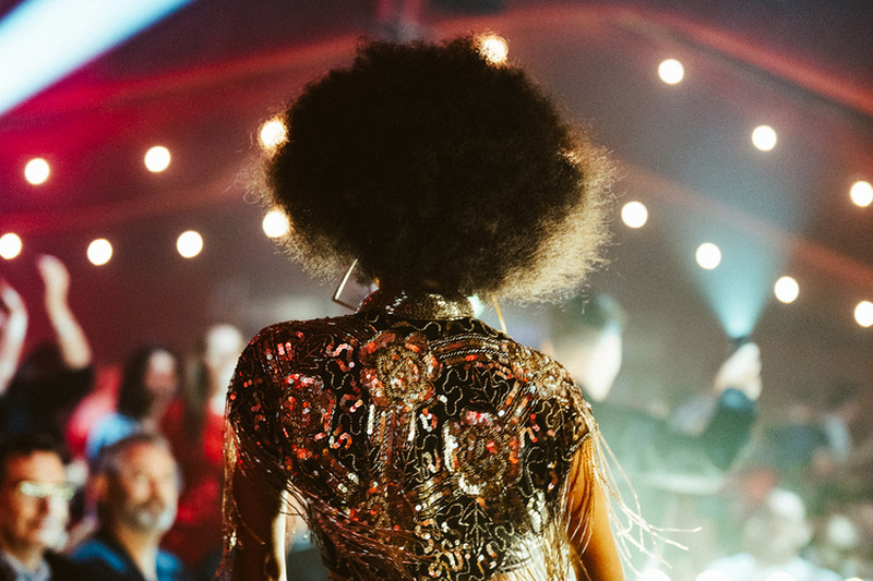 A cabaret performer in a sequinned fringe jacket walking past a seated hens audience