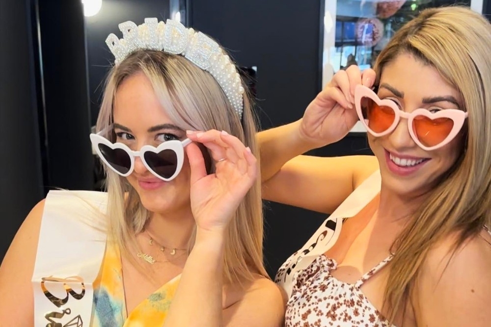 A bride to be in a pearl headband and her friend lowering heart shaped sunglasses