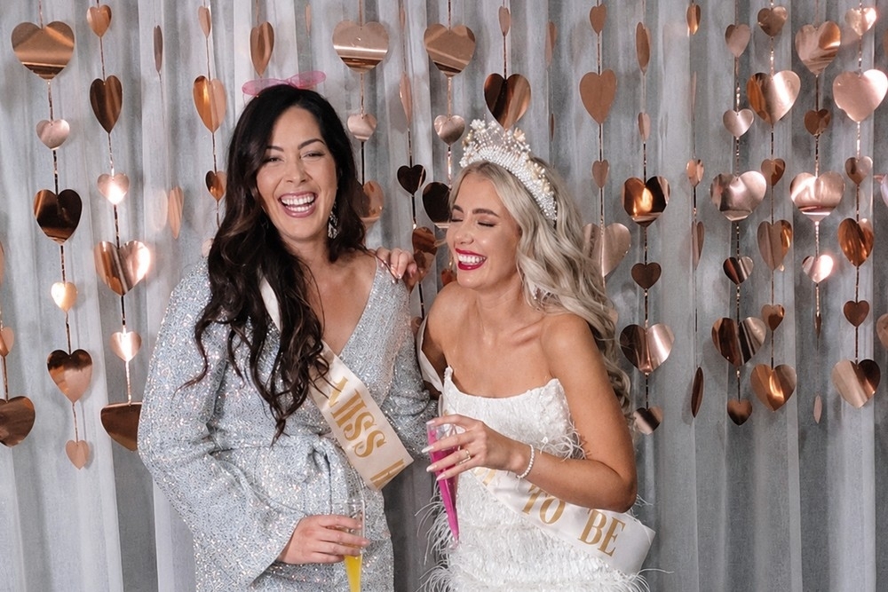 A bride in a pearl crown laughs with a friend in front of a rose gold heart curtain