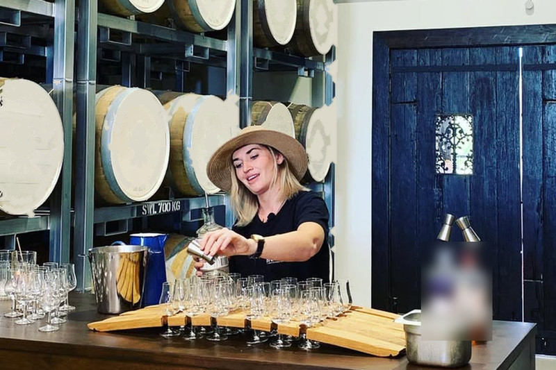 A tasting host in a wide-brimmed hat pouring samples in front of a wall of barrels