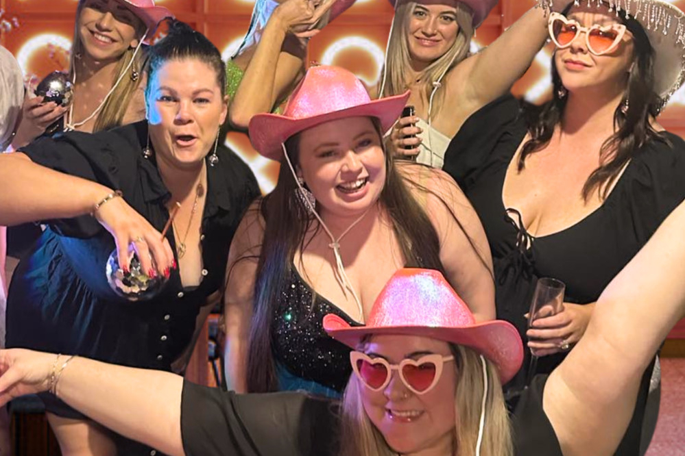 A hens crew in pink cowgirl hats and heart sunglasses cheering at a bar
