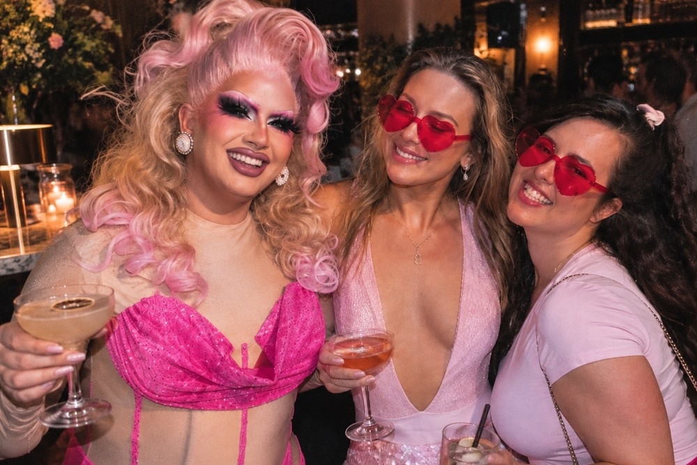 Our drag queen in pink curls and pink sequins raising a cocktail with two hens in red heart sunglasses, all three grinning