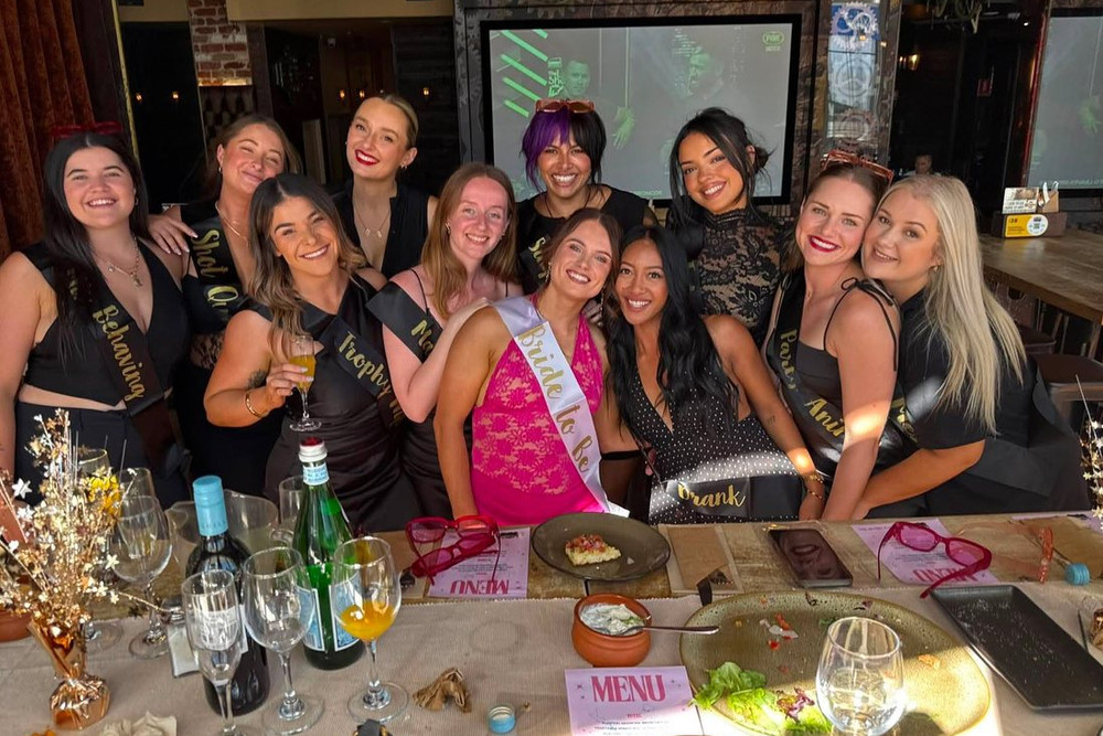 A big hens crew in sashes gathered behind a long table of plates and drinks