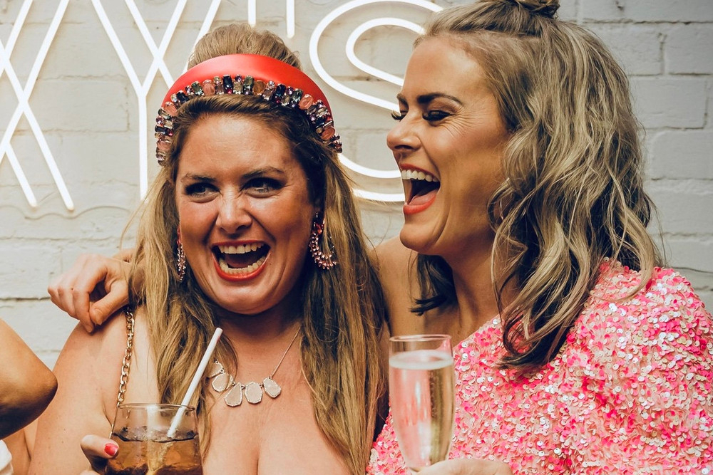 Two friends laughing with drinks in hand, one in a jewelled headband