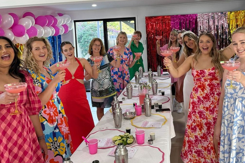 Hens in summer dresses holding pink cocktails around a long table in a living room