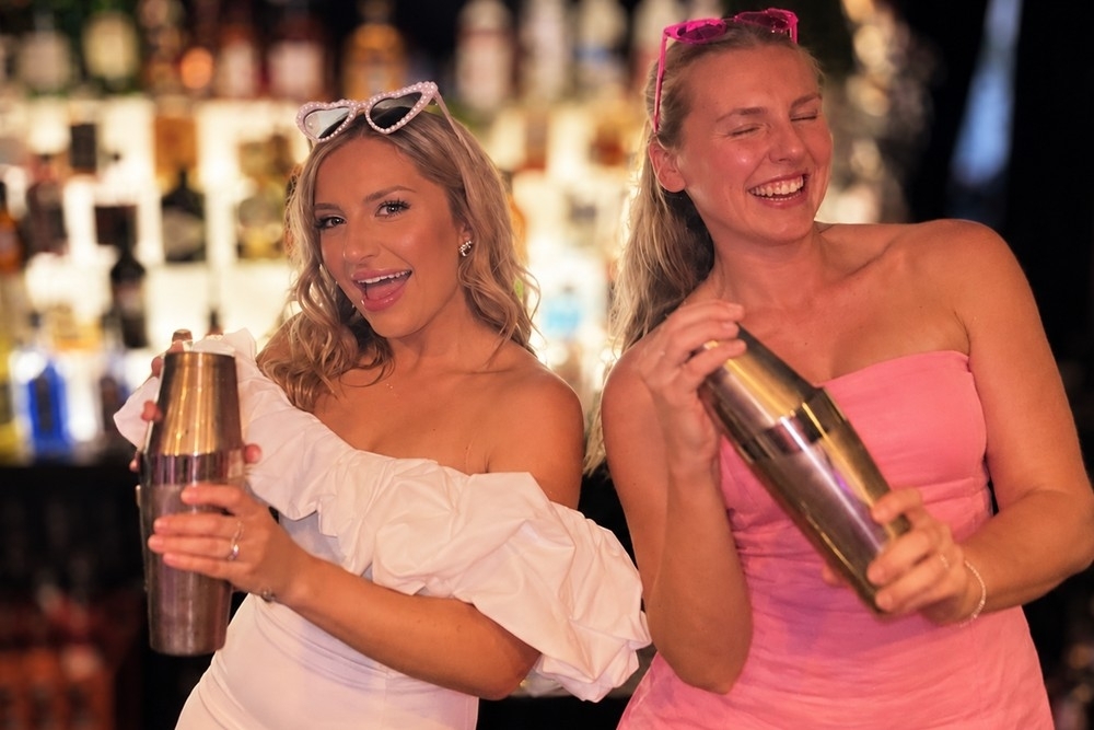 Two friends, one in white and one in pink, laughing as they shake cocktails in front of a glowing back bar
