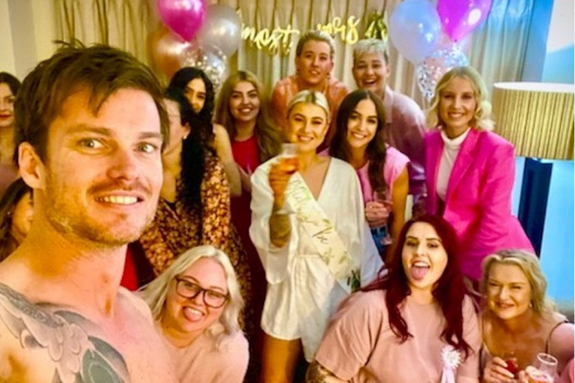 A shirtless performer taking a selfie with a laughing hens crew and pink balloons