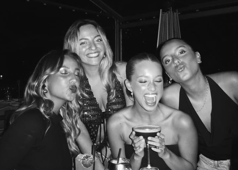 Four friends pulling faces and laughing over cocktails at a dinner table, in black and white