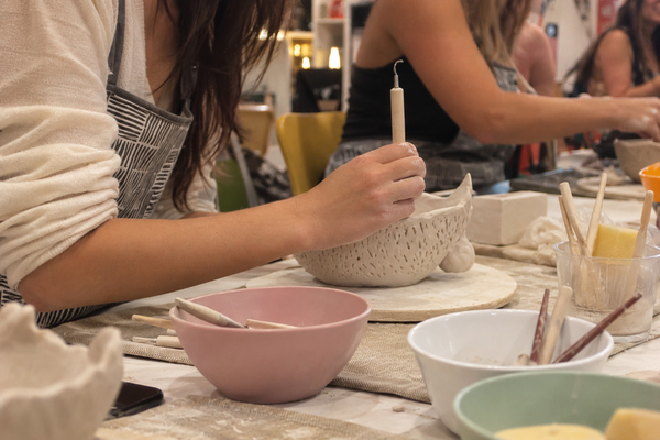 Hands shaping a textured clay bowl beside pastel mixing bowls and tools