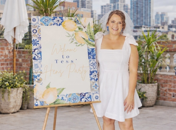 A smiling bride in a short white dress and veil beside a lemon and blue tile hens party welcome sign on a sunny rooftop