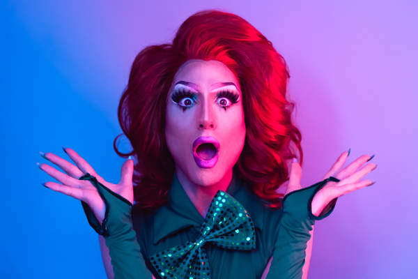 A drag queen in a red wig and sequin bow tie, mouth open in mock shock
