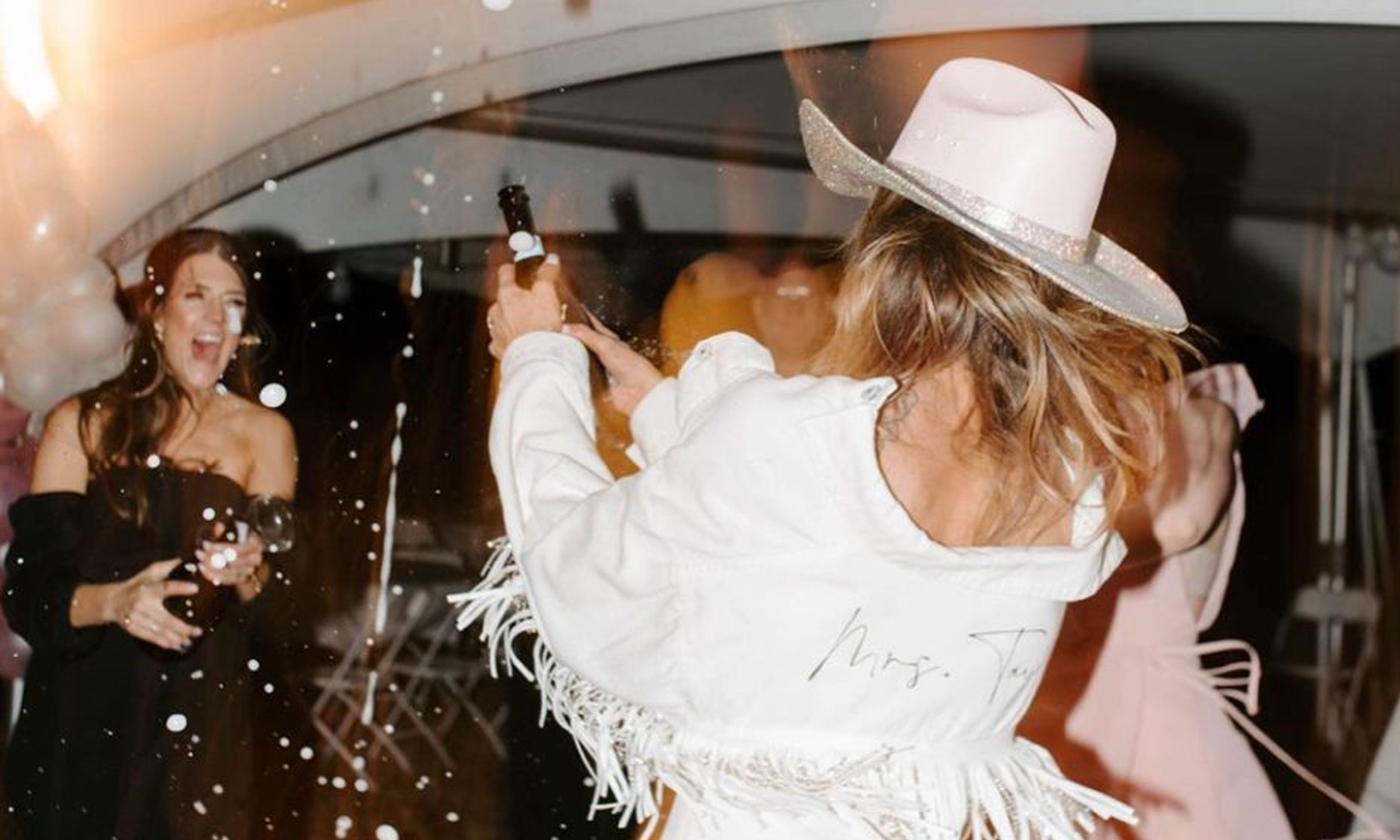 Friends spraying champagne in celebration, one in a white fringe jacket and hat